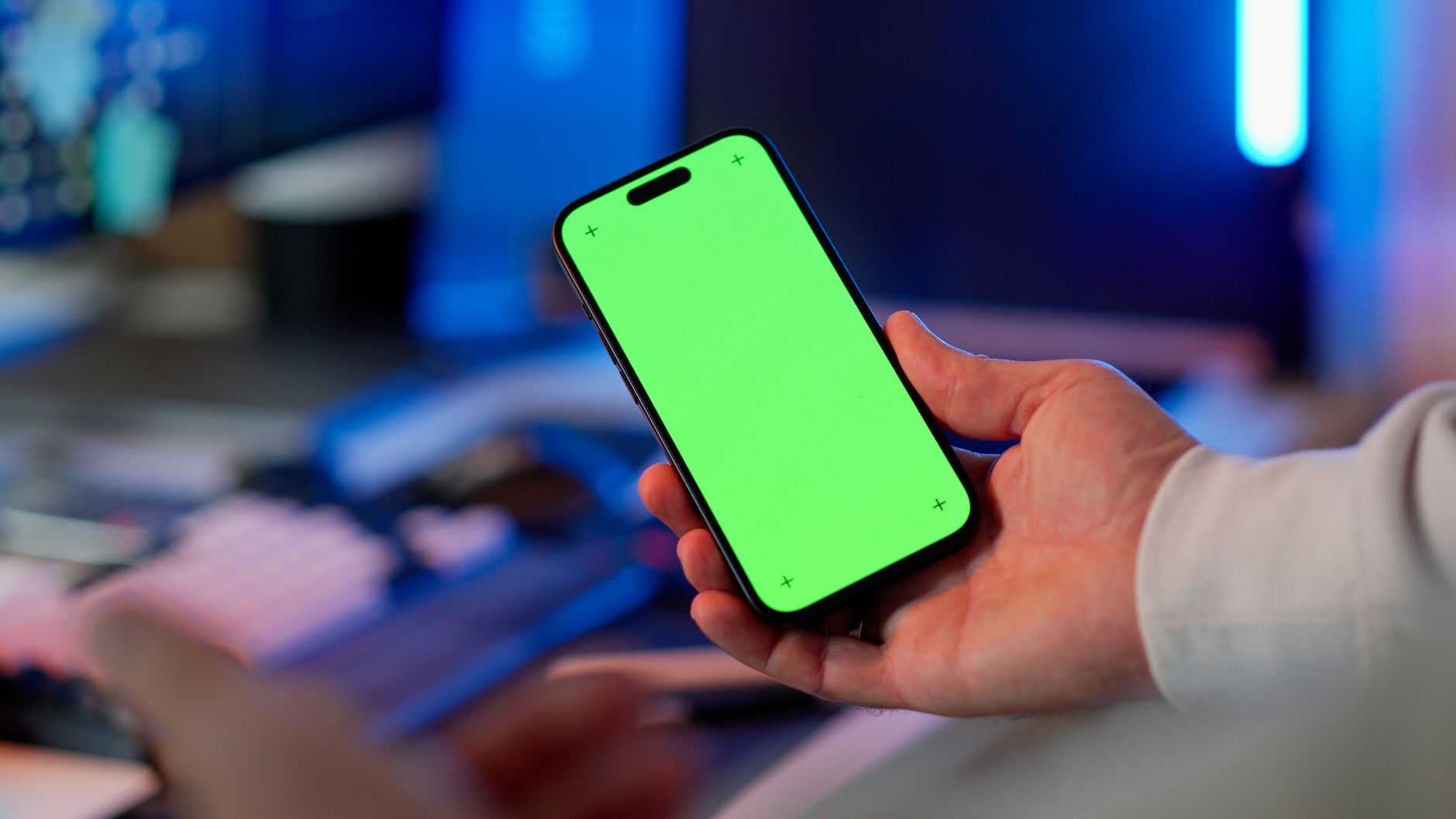 Une main tient un smartphone à l’écran vert devant un bureau éclairé de néons bleus.